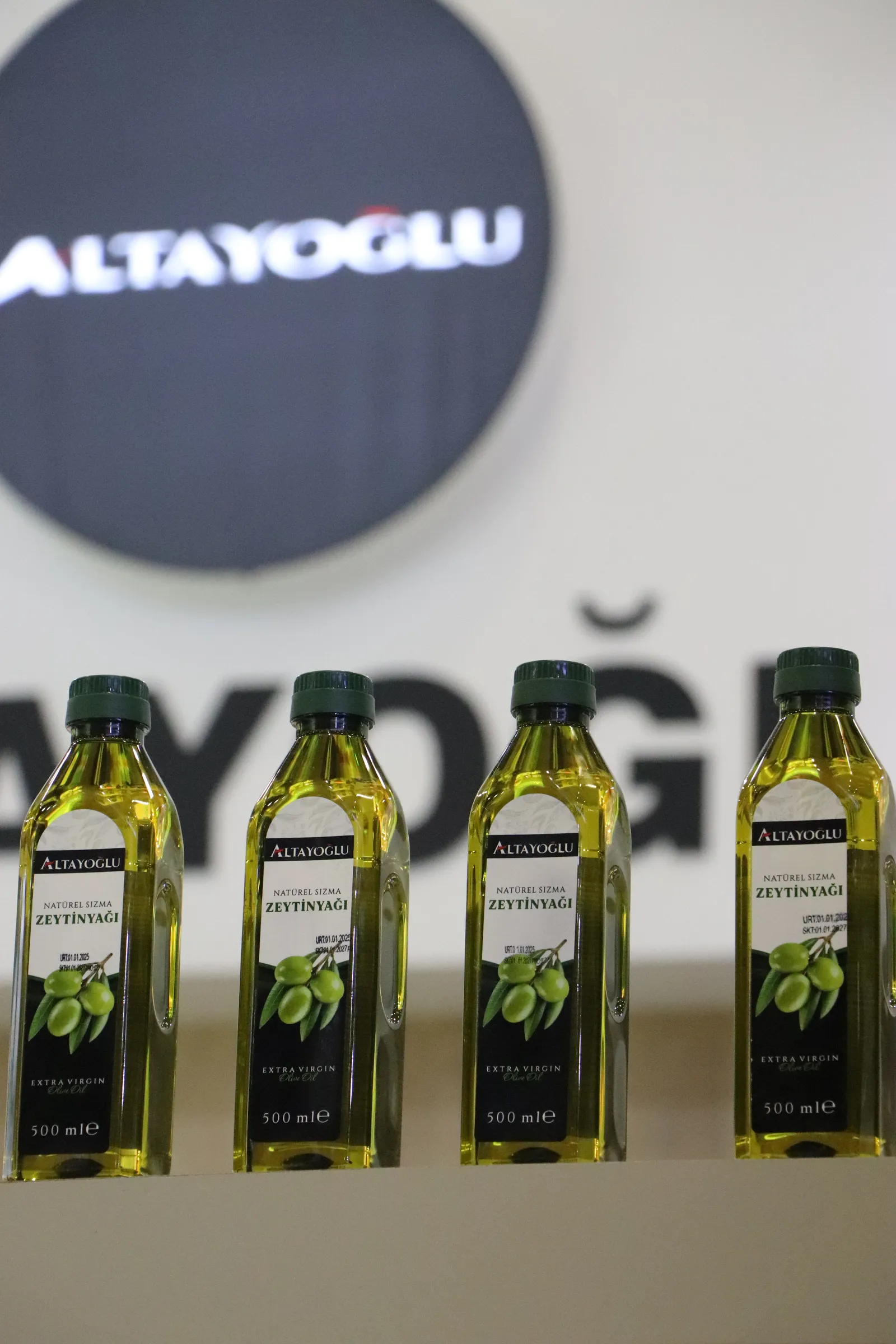 Fuar standında sergilenen zeytinyağı şişeleri, arka planda bulanık marka logosu