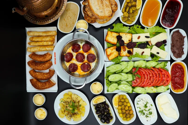 Koyu masada serpme kahvaltı: sucuklu yumurta, peynirler, zeytin, domates, salatalık ve börek; üstten